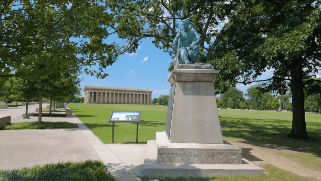 View of Nashville's Parthenon from across the lawn in Centennial Park on a sunny day, with a statue in the foreground and the full-scale Greek temple replica visible in the distance surrounded by green trees and open space