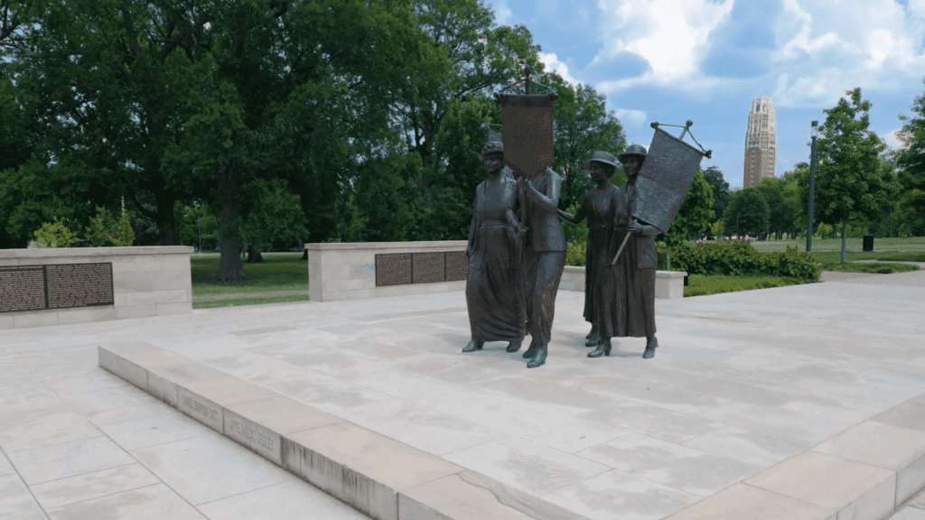 Tennessee Woman Suffrage Monument in Centennial Park featuring bronze statues of women suffragists with protest signs, with the Parthenon visible in the background among trees on a sunny day.