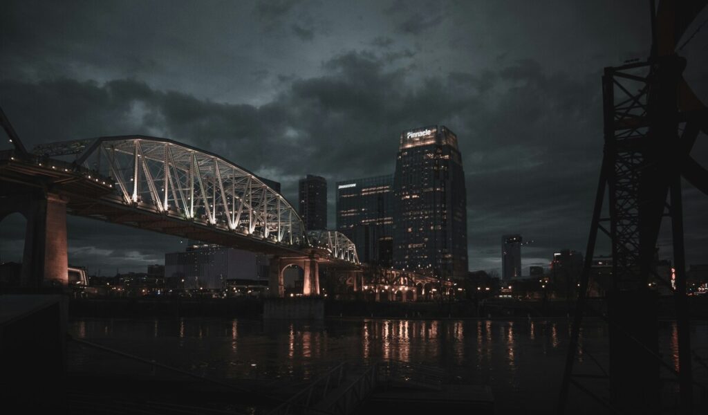 Illuminated steel bridge with arch design spanning river at night, with city skyline, Pinnacle building, and dramatic stormy sky