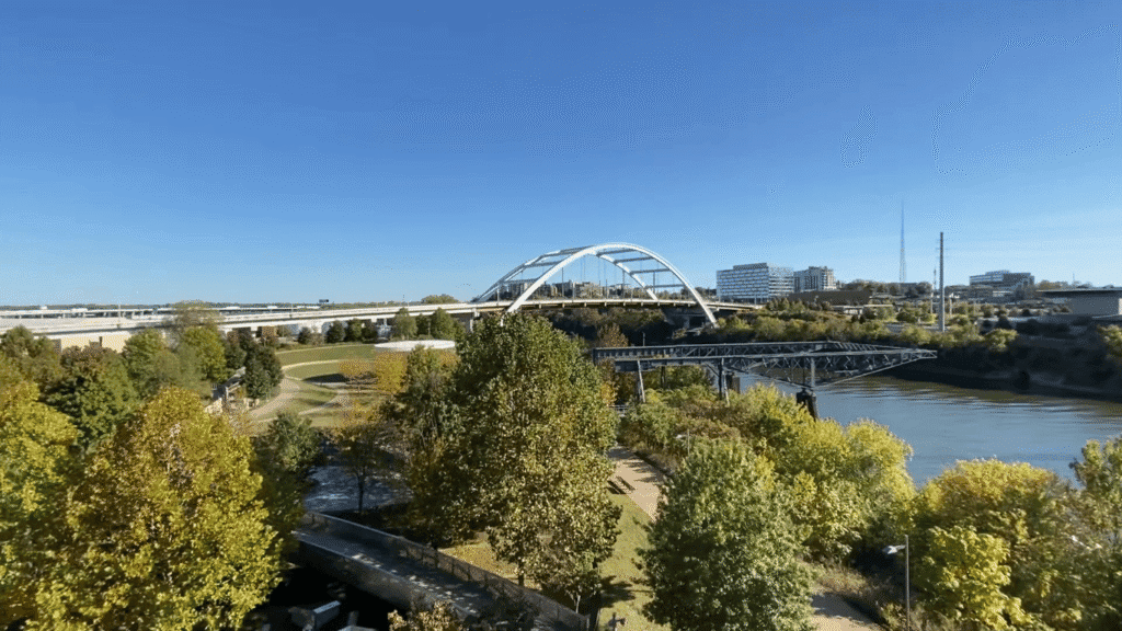  White arch bridge spanning river with parkland and walking paths below, city buildings visible in background under clear blue sky