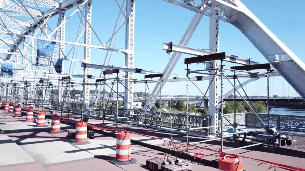  Steel bridge structure under construction with temporary scaffolding, orange traffic barriers, and white metal framework spanning across a river