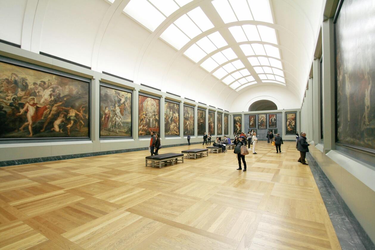 Spacious art gallery with high arched ceiling, wooden floors, and classic paintings displayed on white walls