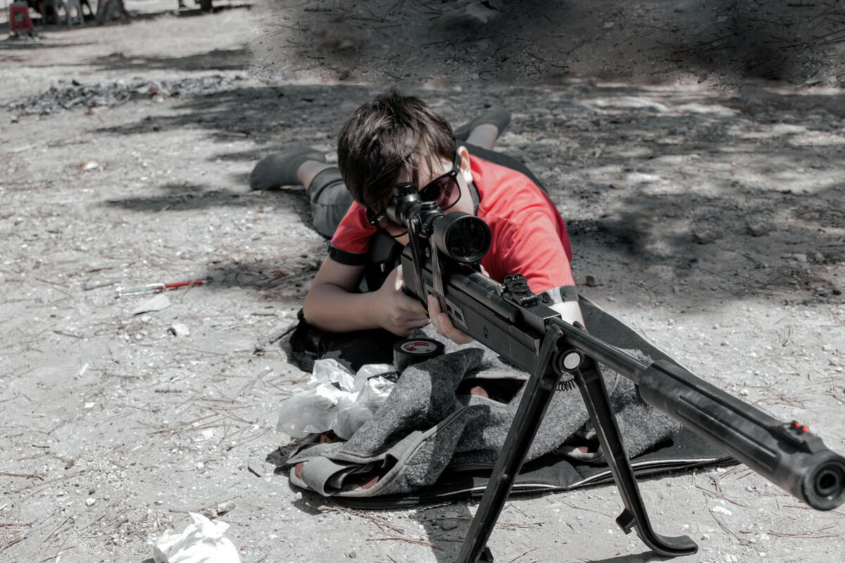 Young person in red shirt lying prone aiming with scoped rifle at outdoor shooting range Nashville