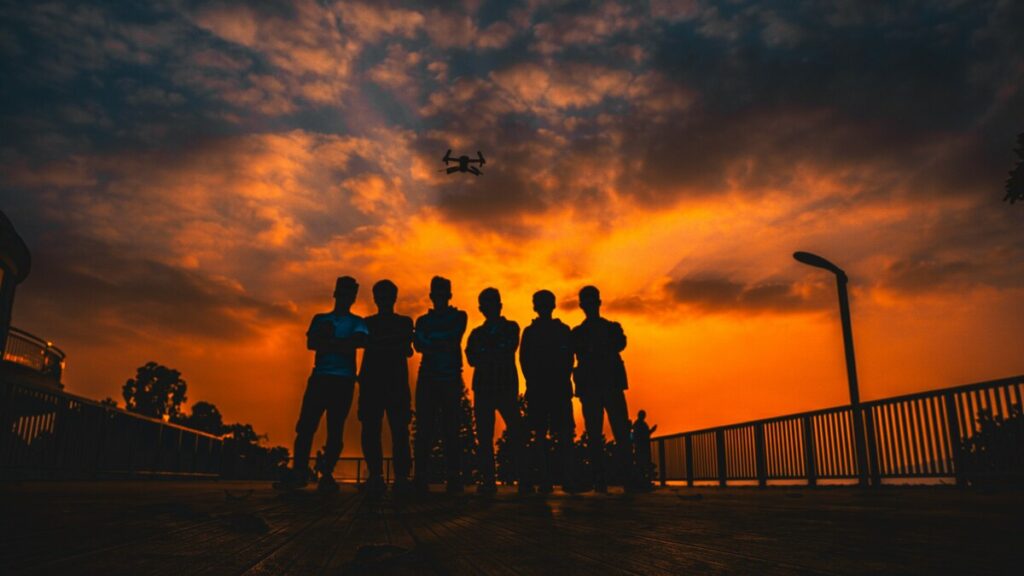 Silhouettes of friends standing together on a wooden deck against a dramatic orange sunset sky with a drone flying overhead