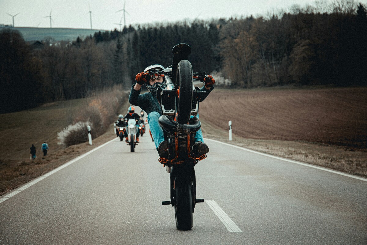 A motorcyclist pops a wheelie on an open country road, riders trailing behind, wind turbines dotting the distance