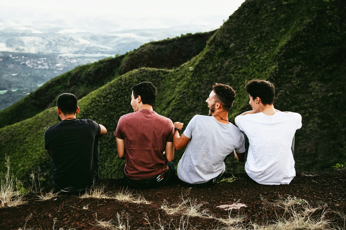 Four friends sitting side by side on a mountain overlook, enjoying a panoramic view of green valleys and distant hills